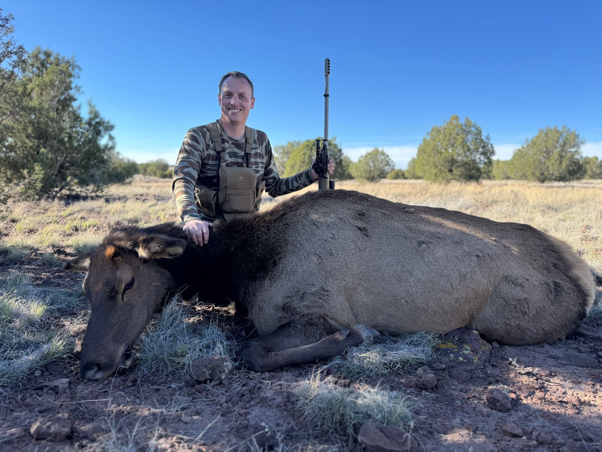 Person smiles, stands over an elk lying on ground, holding a rifle in a field.