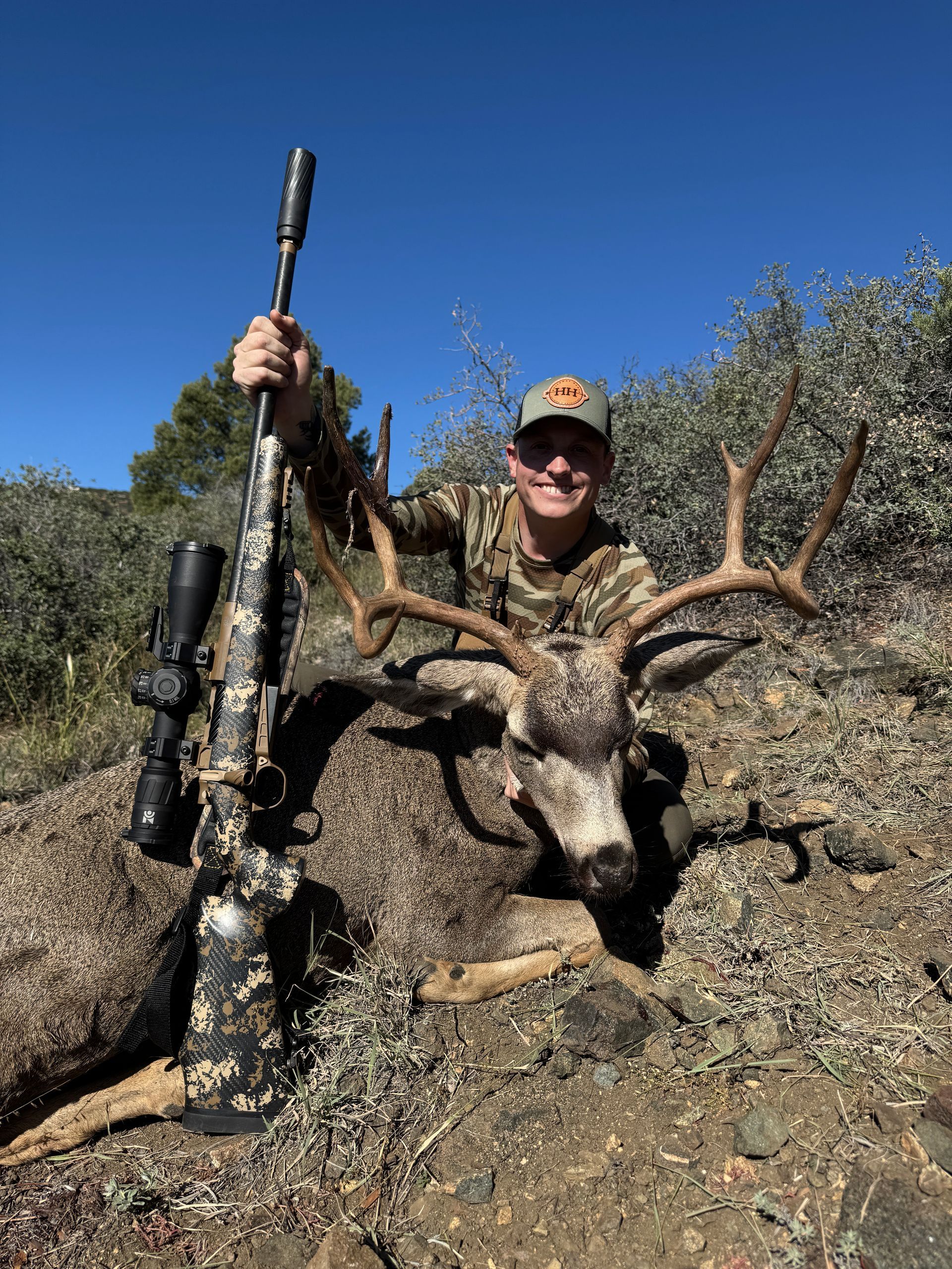 Hunter smiles, holding rifle, posing next to large deer with antlers in sunny outdoor setting.