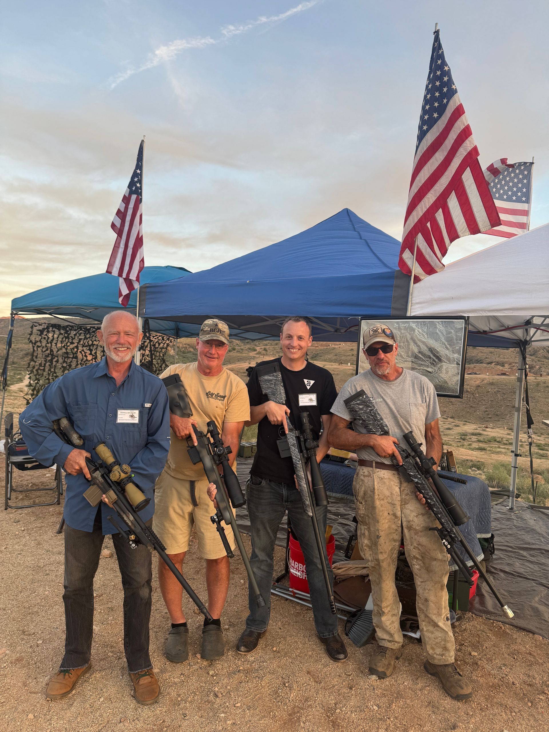 Four men pose with rifles under a tent with American flags in a desert setting.
