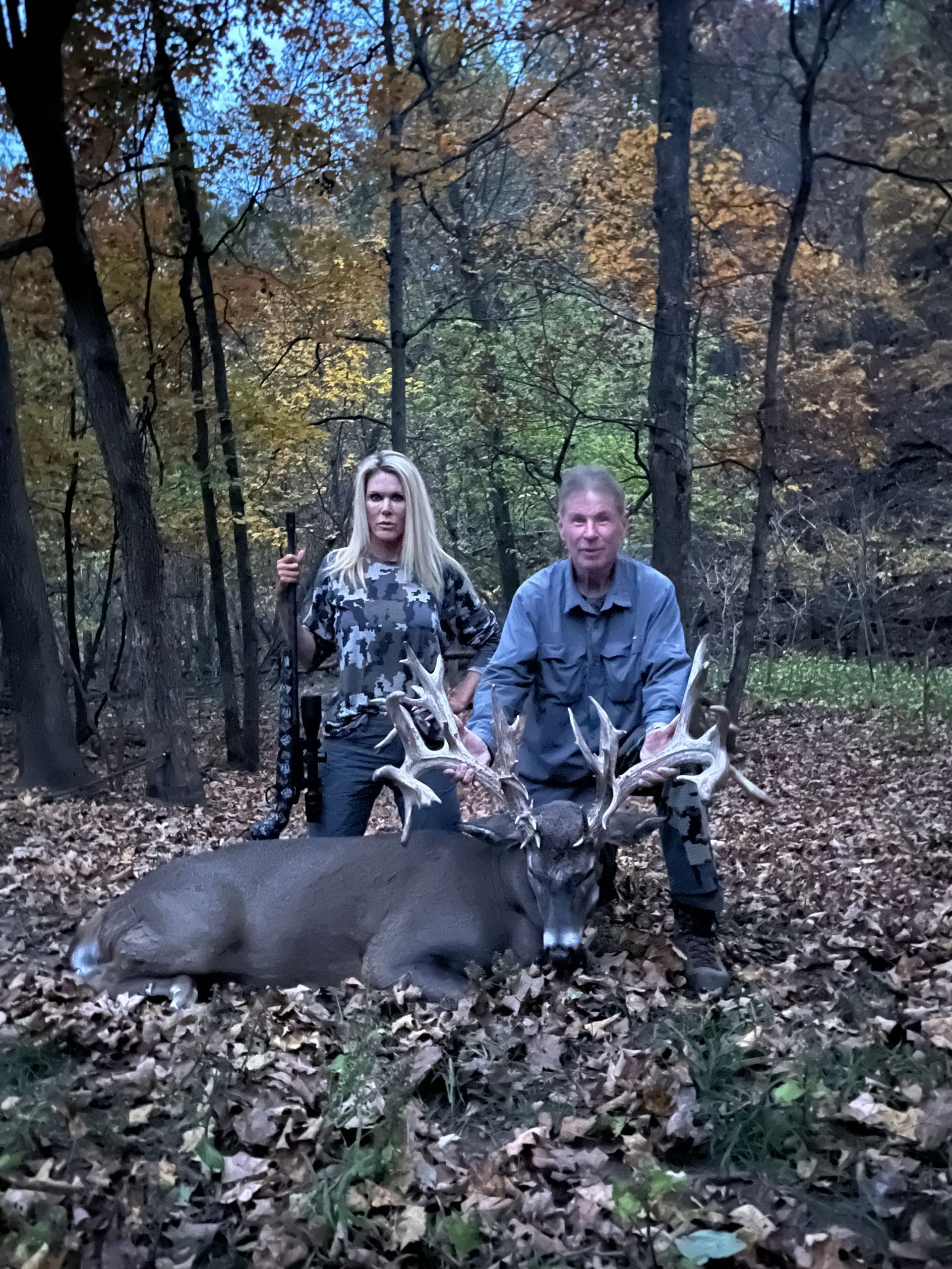 A person and a person posing with a large dead deer in a forest, both smiling. The person is holding a rifle.