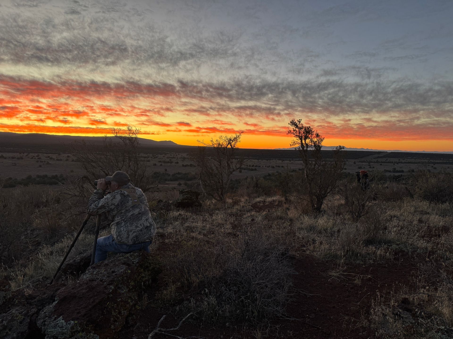 Photographer kneels, tripod set, capturing sunrise. Orange and yellow sky over open field, dark silhouettes of shrubs.