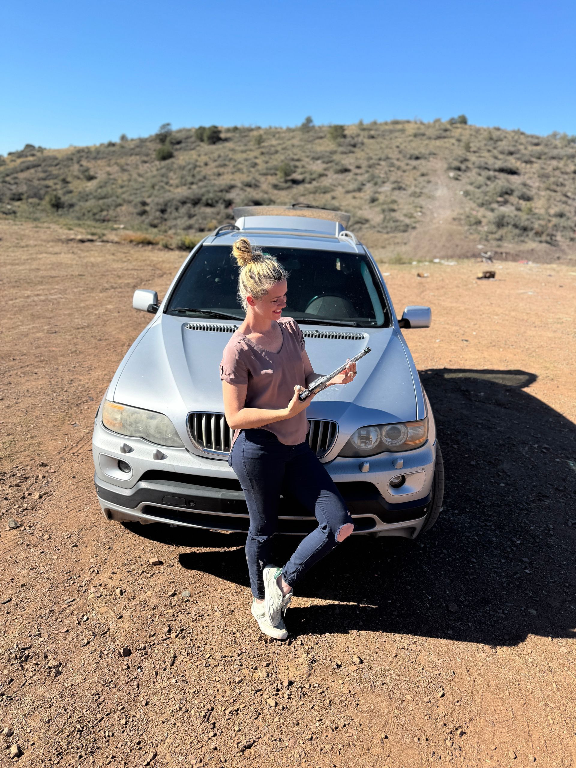 Woman holding a stick poses in front of a silver SUV in a desert setting on a sunny day.