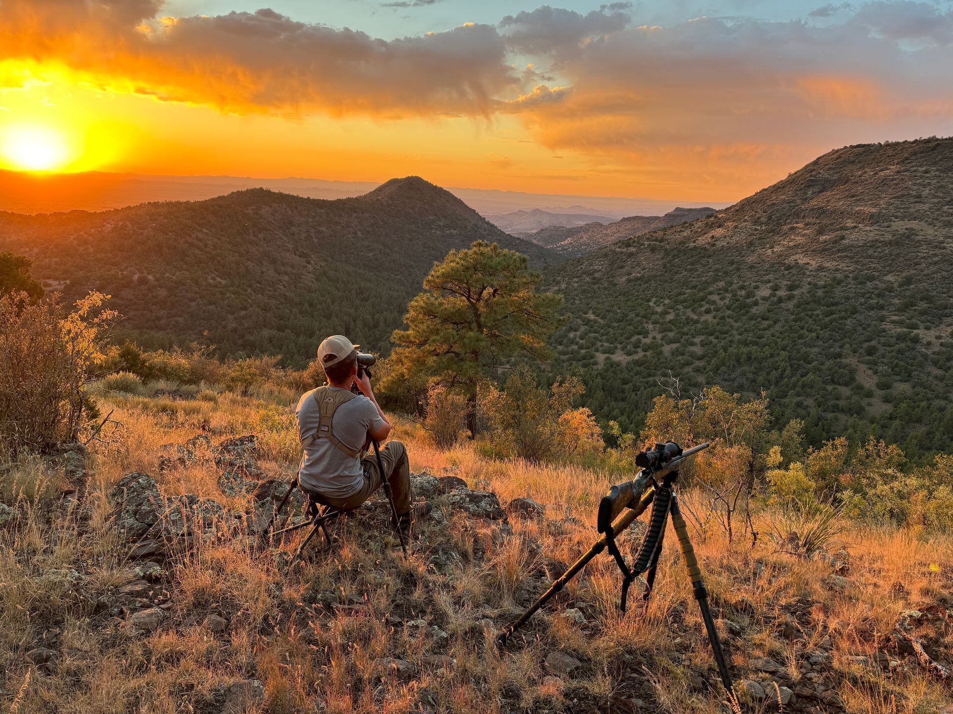 Man with binoculars on tripod overlooking mountains at sunset.