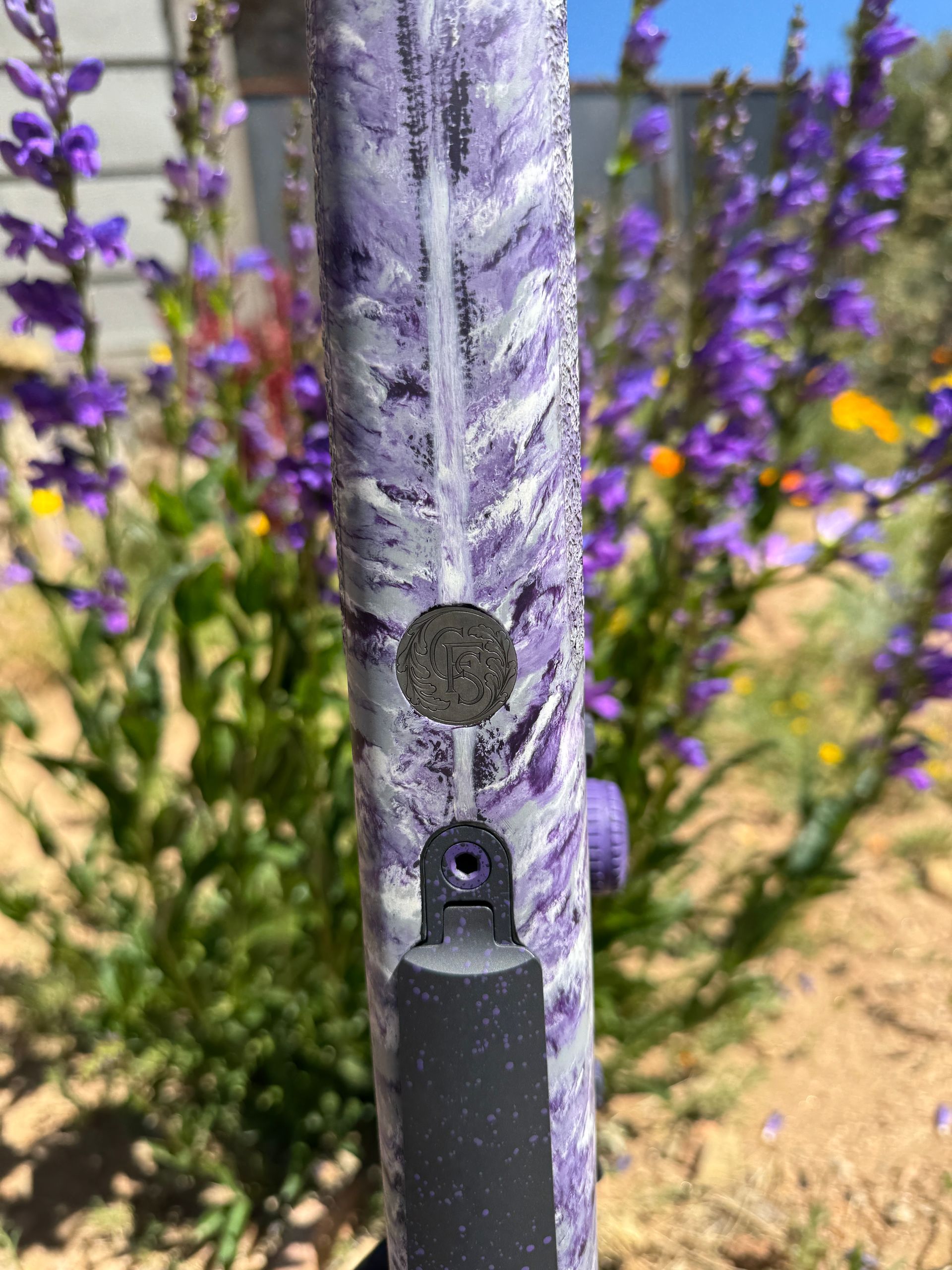 Close-up of a purple and white camouflage rifle stock, set against a background of purple flowers.