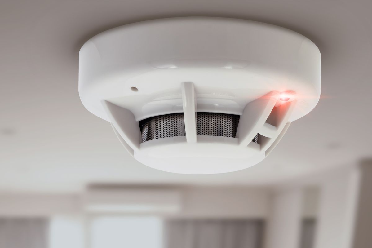White smoke detector on a ceiling with a red light illuminated, in a room setting.