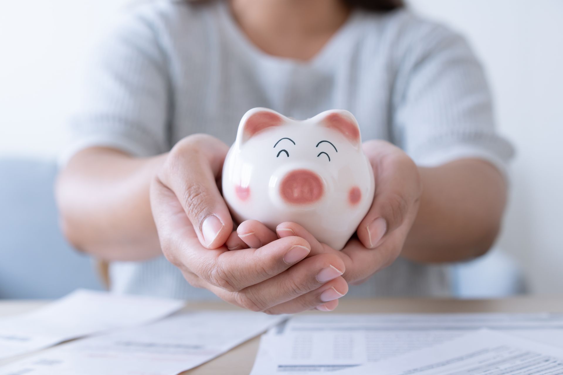 Hands holding a smiling piggy bank, papers on the table.