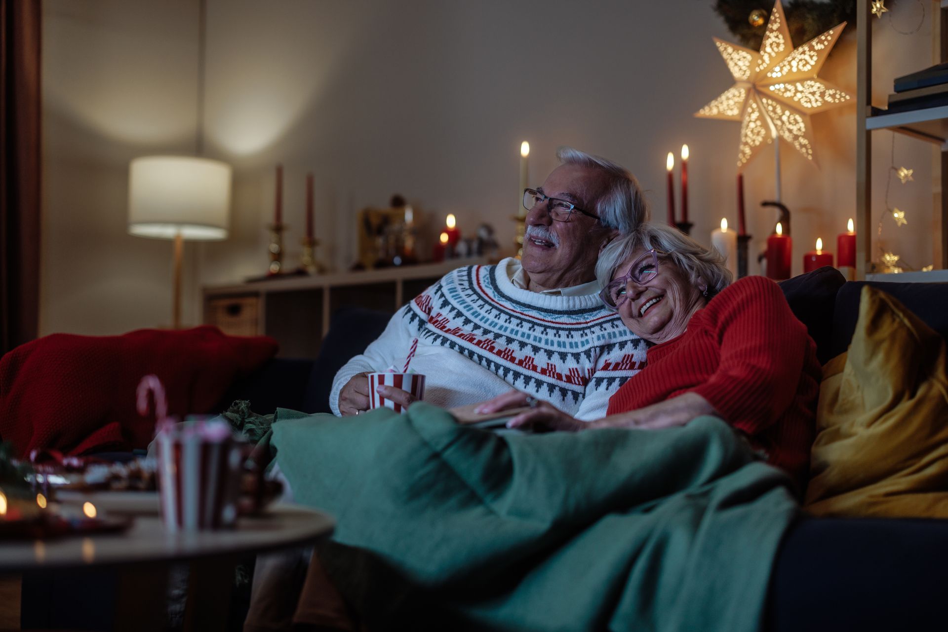Elderly couple cuddled on a couch watching TV in a cozy, Christmas-decorated living room.