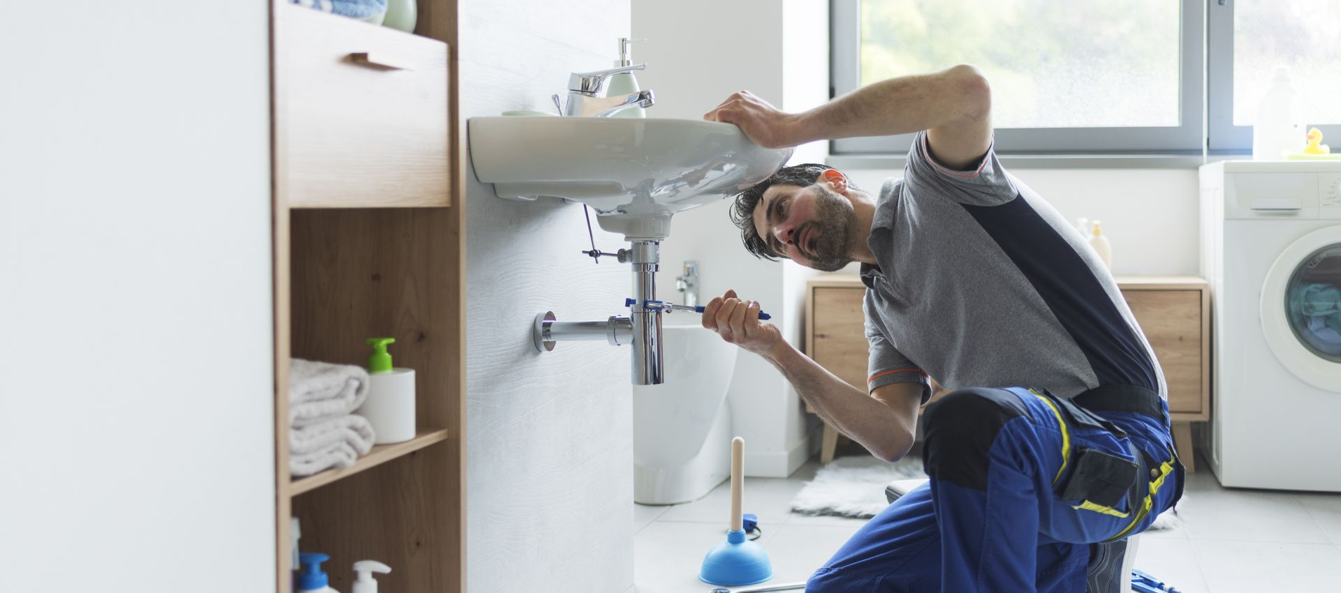 Plumber kneeling, fixing sink in bathroom. Blue overalls, gray shirt, tools on floor.
