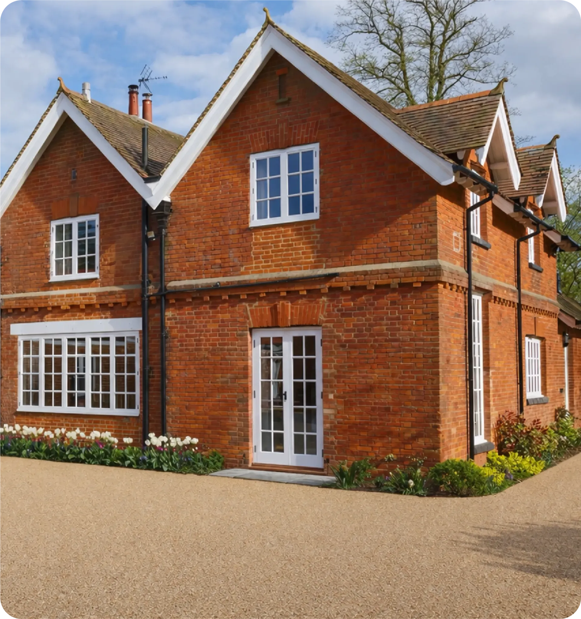 A two-story red brick house with white window frames and a white double door, set behind a light-colored gravel driveway.