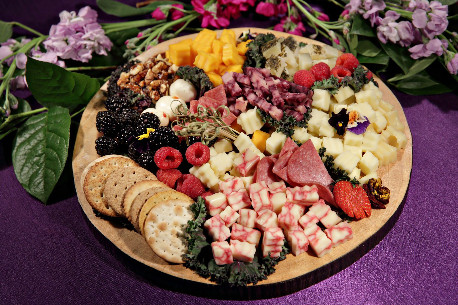 A wooden cutting board filled with different types of food and crackers on a purple table.