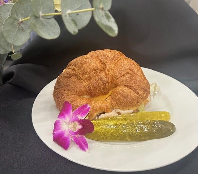 A croissant sandwich with pickles and a purple flower on a white plate