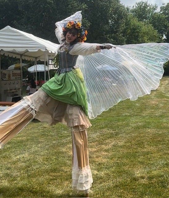 A woman in a green dress and wings is standing on stilts in a field.