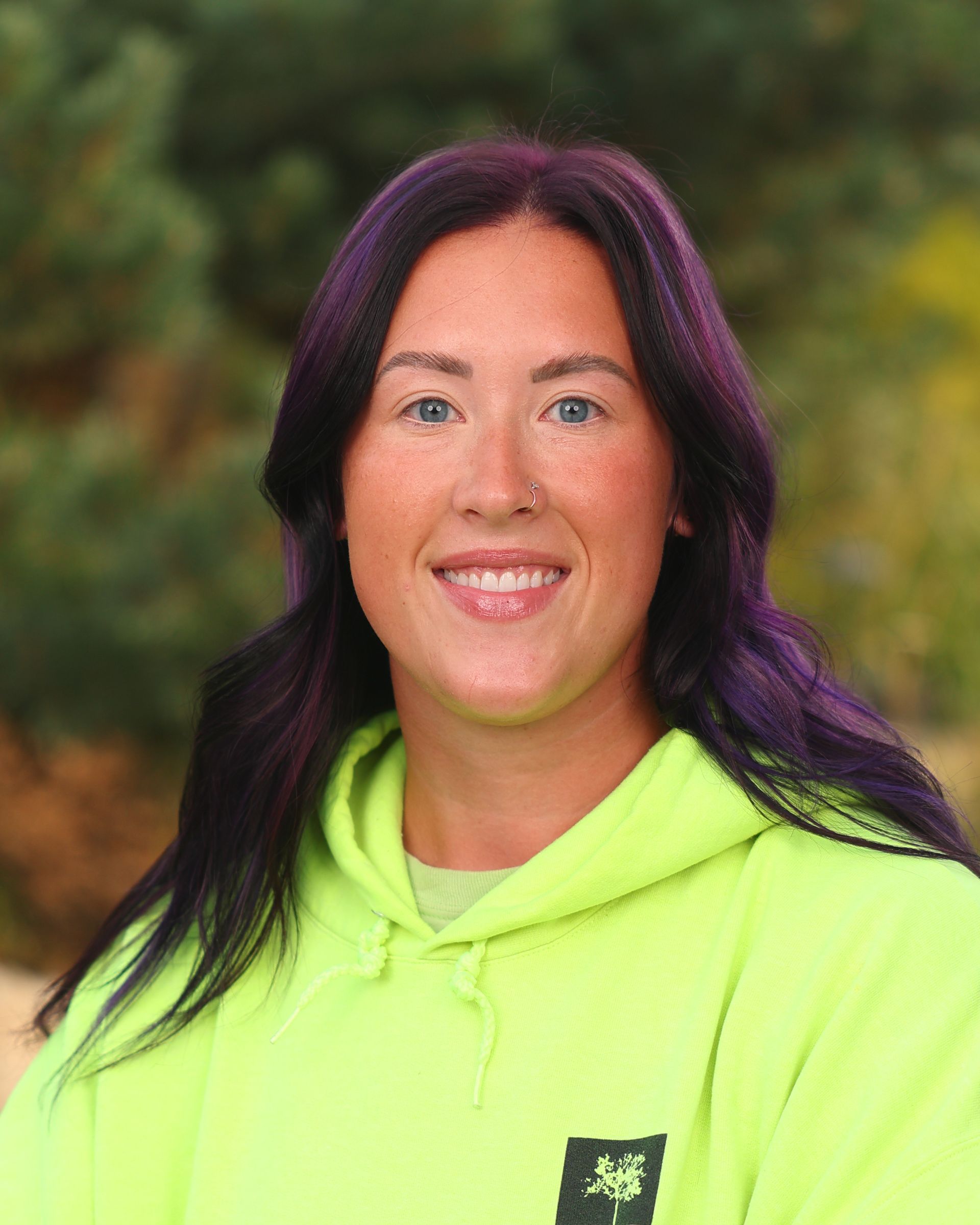 A woman with purple hair is wearing a yellow hoodie and smiling.