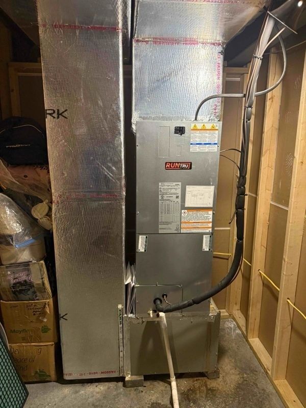 Furnace in a utility room with ductwork. Metal and silver unit against wood-framed walls and concrete floor.