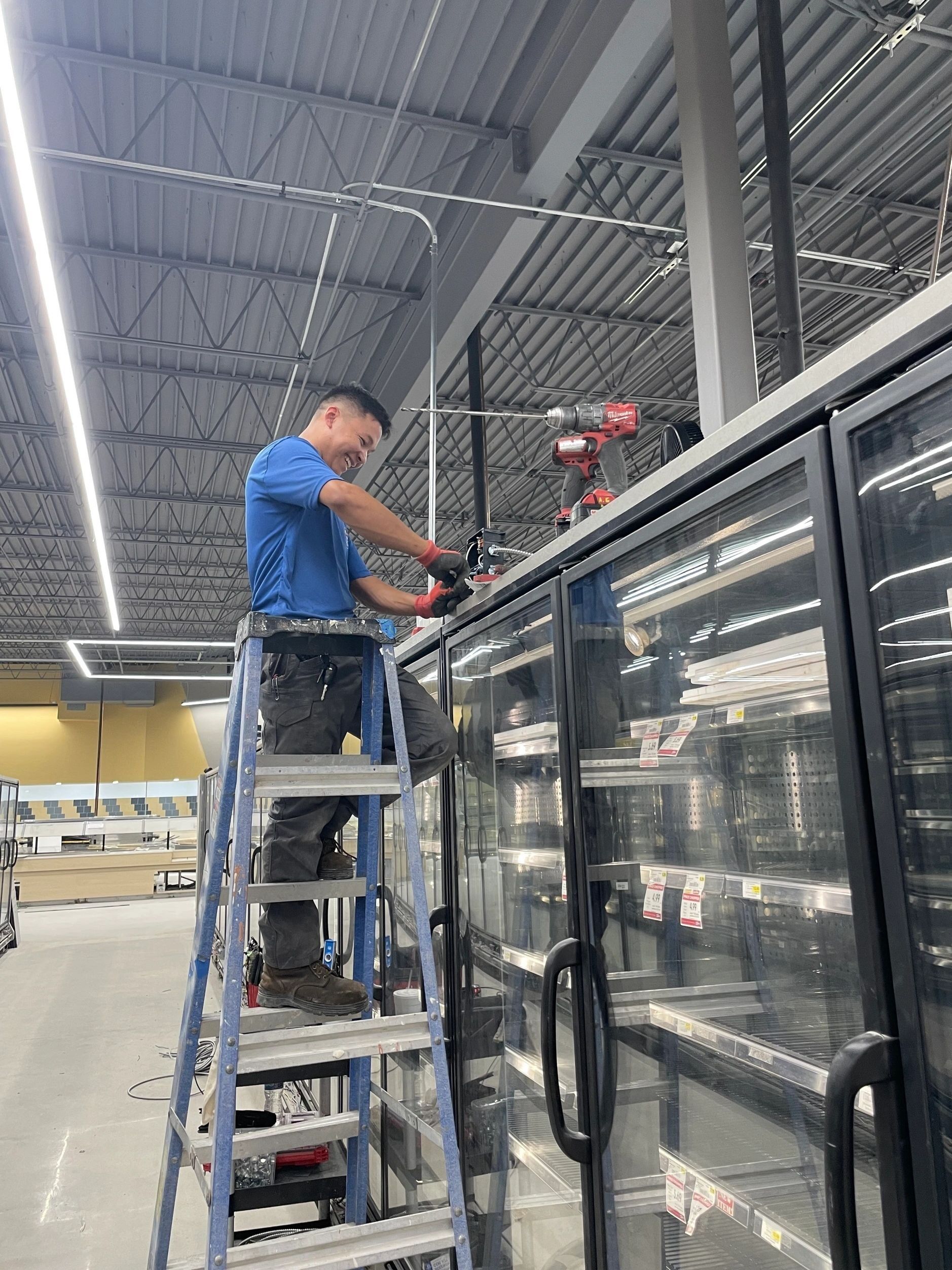 Man in blue shirt uses a drill on top of a refrigerator, standing on a ladder in a store.
