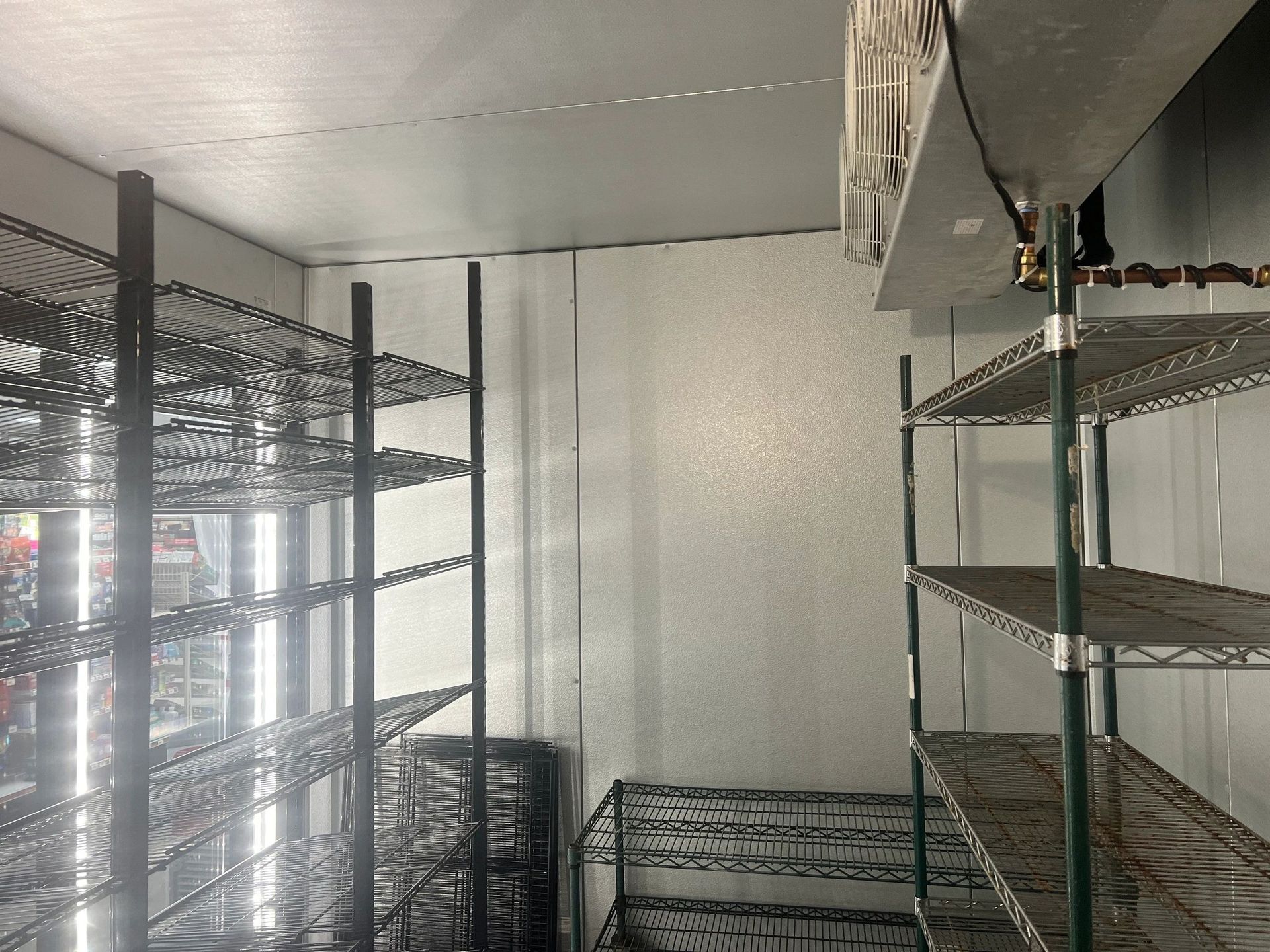 Empty commercial walk-in cooler with wire shelving; white walls, metal ceiling, and visible refrigeration unit.