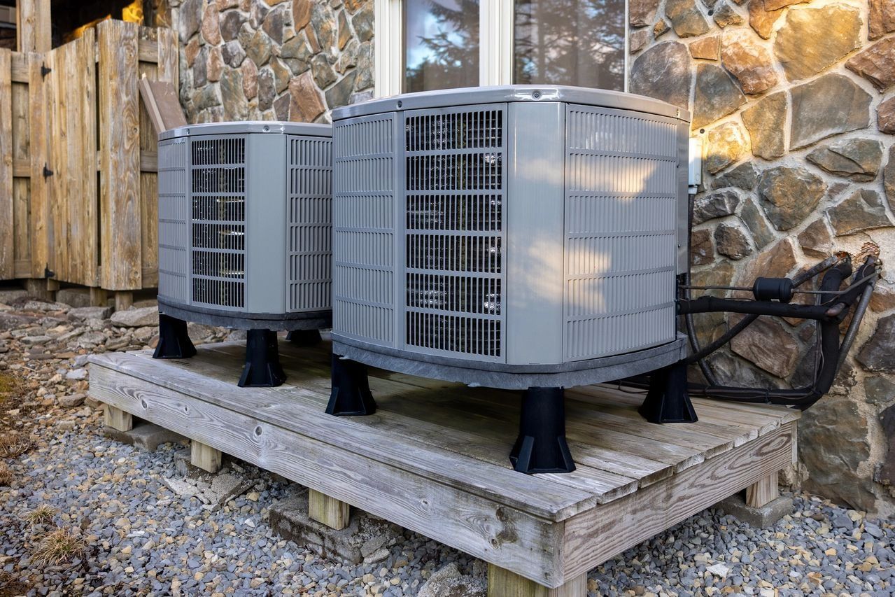 Two gray air conditioning units on a wooden platform near a stone wall.