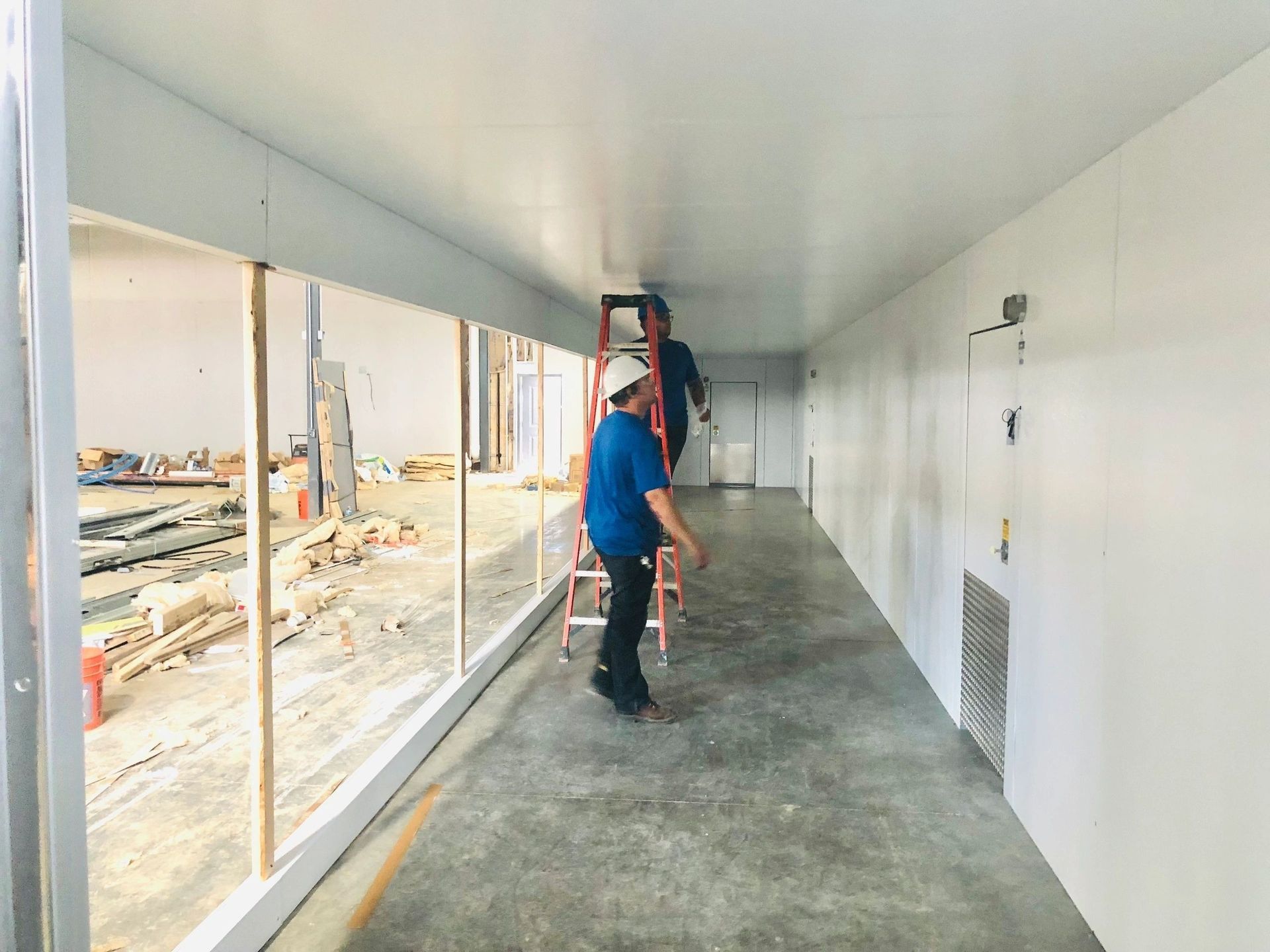 Two workers inside a white-walled construction corridor; one on a ladder.