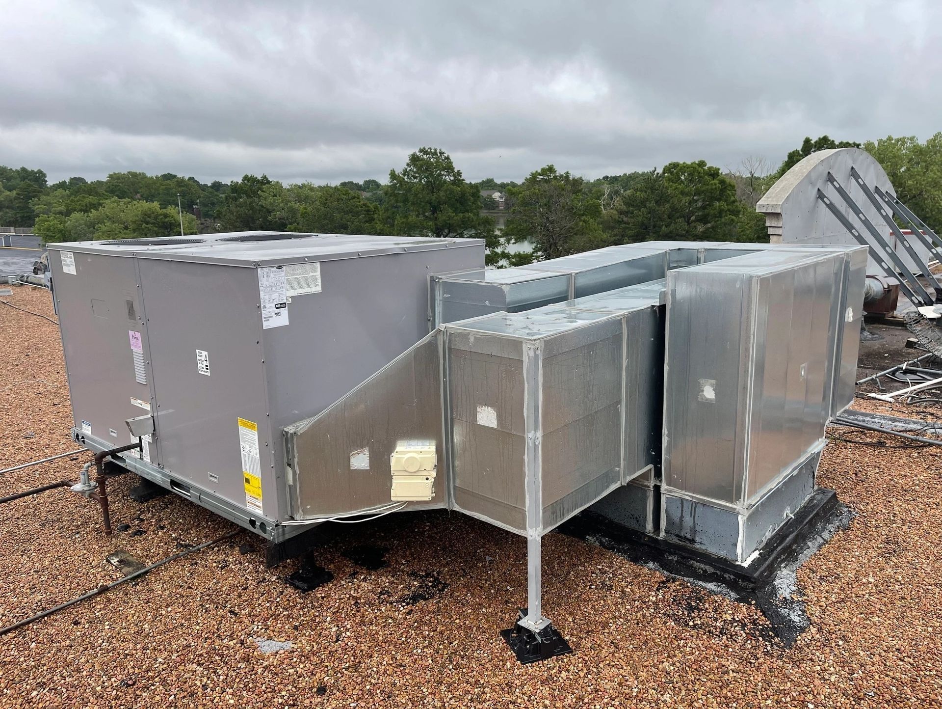 Rooftop HVAC unit, gray metal, connected to ductwork, sitting on a gravel roof under a cloudy sky.