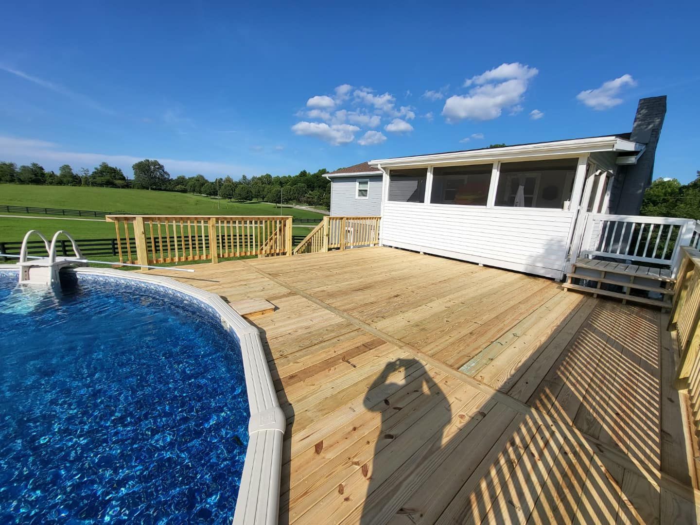 A wooden deck next to a swimming pool with a house in the background.