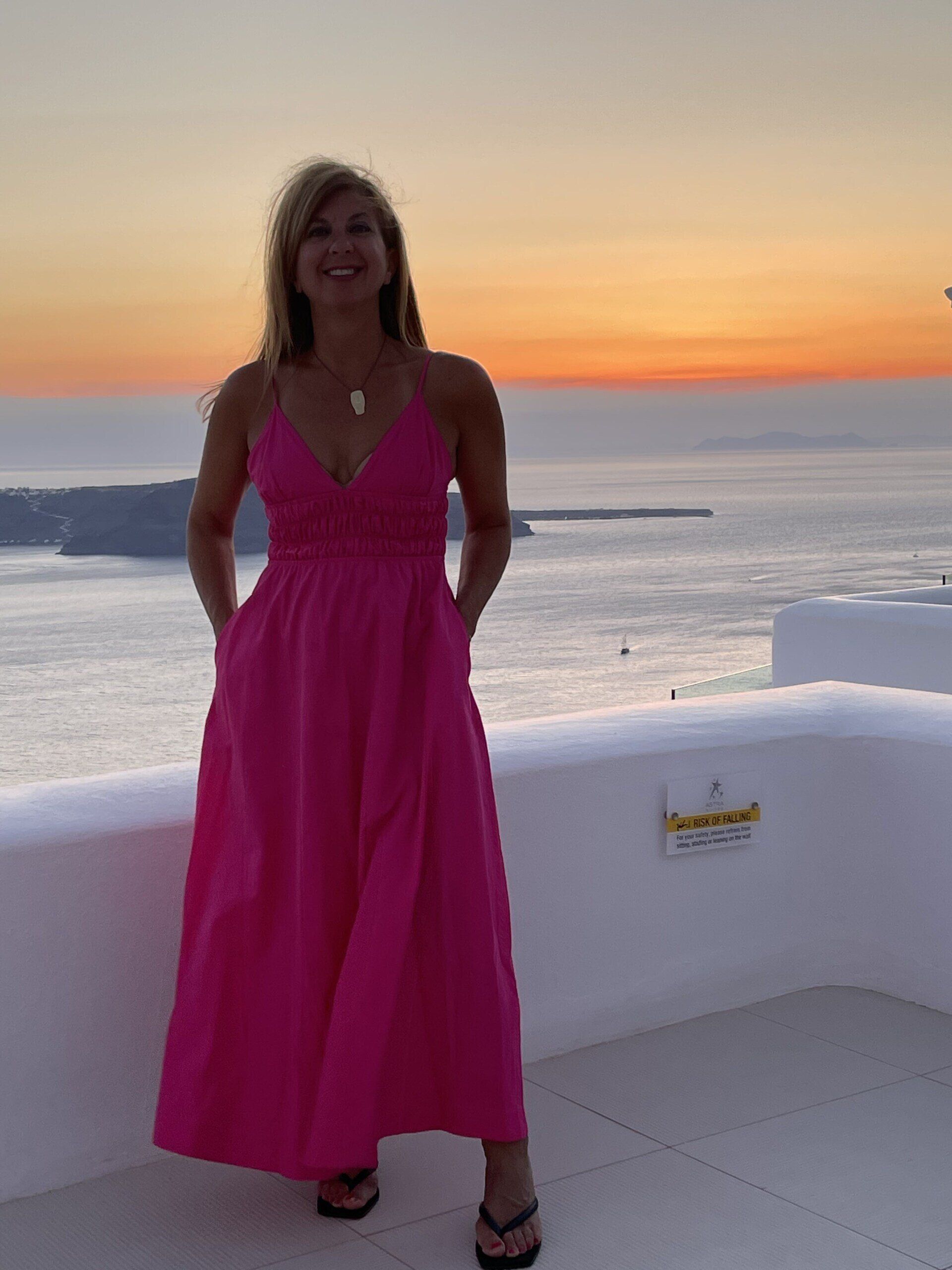 A woman in a pink dress is standing on a balcony overlooking the ocean.