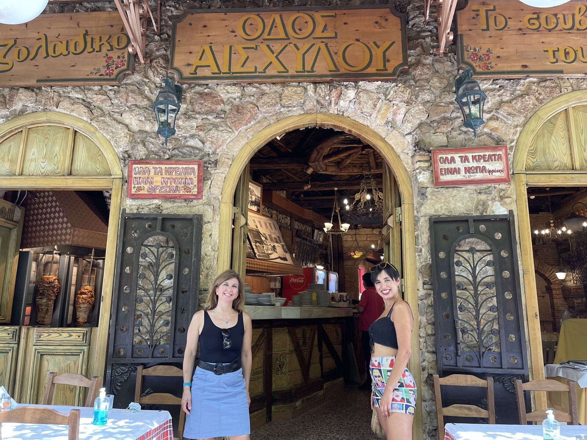 Two women are standing in front of a restaurant with tables and chairs.