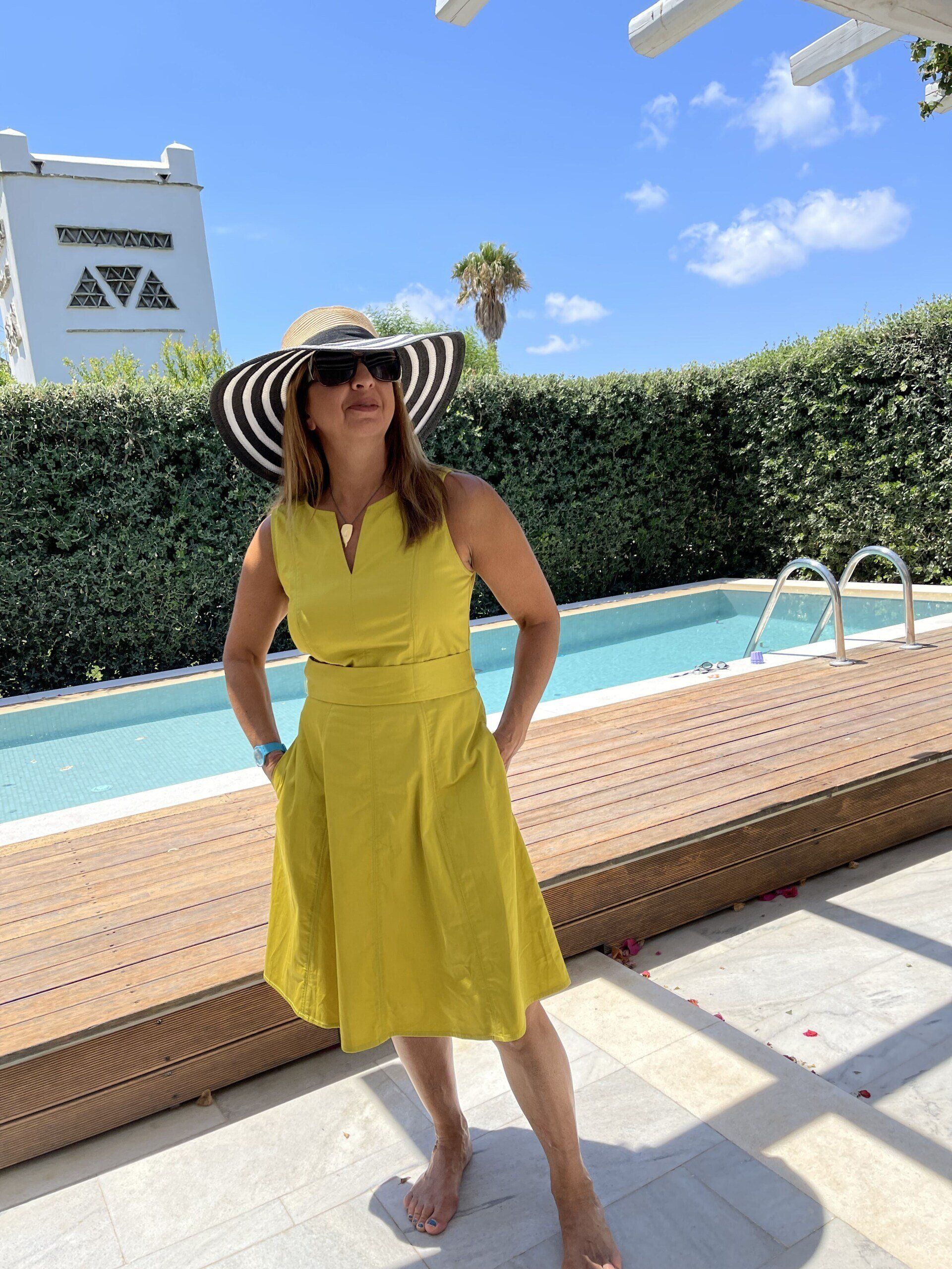 A woman in a yellow dress and hat is standing next to a pool.