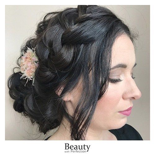 A woman is wearing her hair in a braided bun with a flower in her hair and professional makeup.