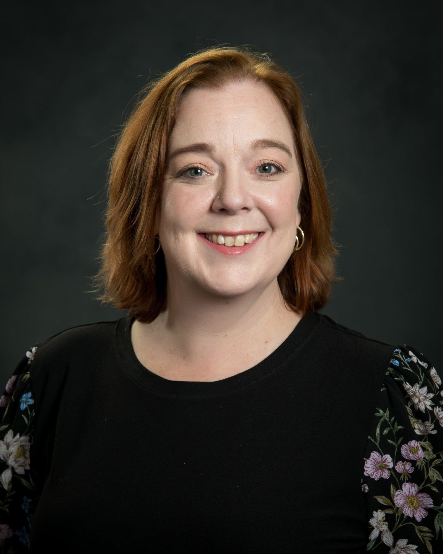 A woman with red hair is smiling and wearing a black shirt with floral sleeves.