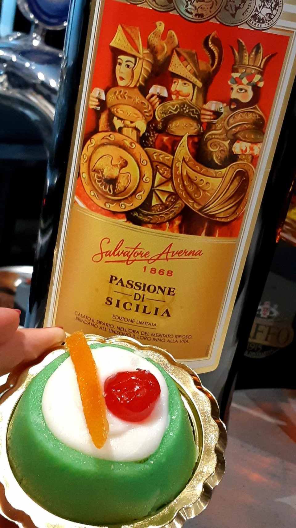 Una persona tiene in mano una cassata davanti a una bottiglia di passione di sicilia