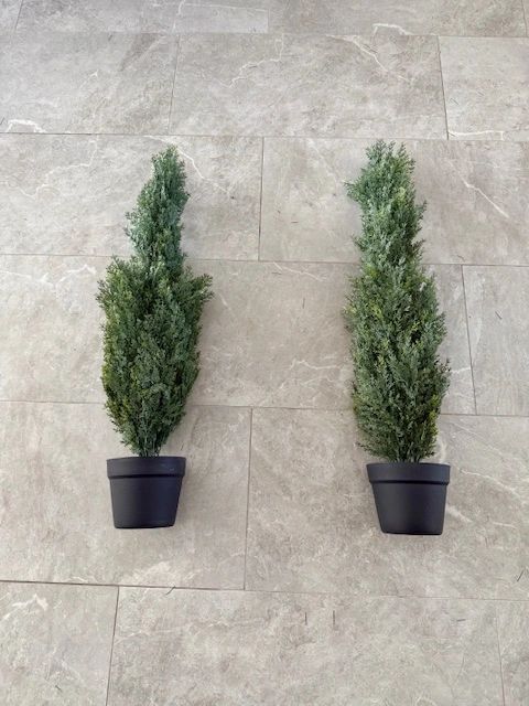 Two potted, artificial cypress trees against a stone tiled wall.