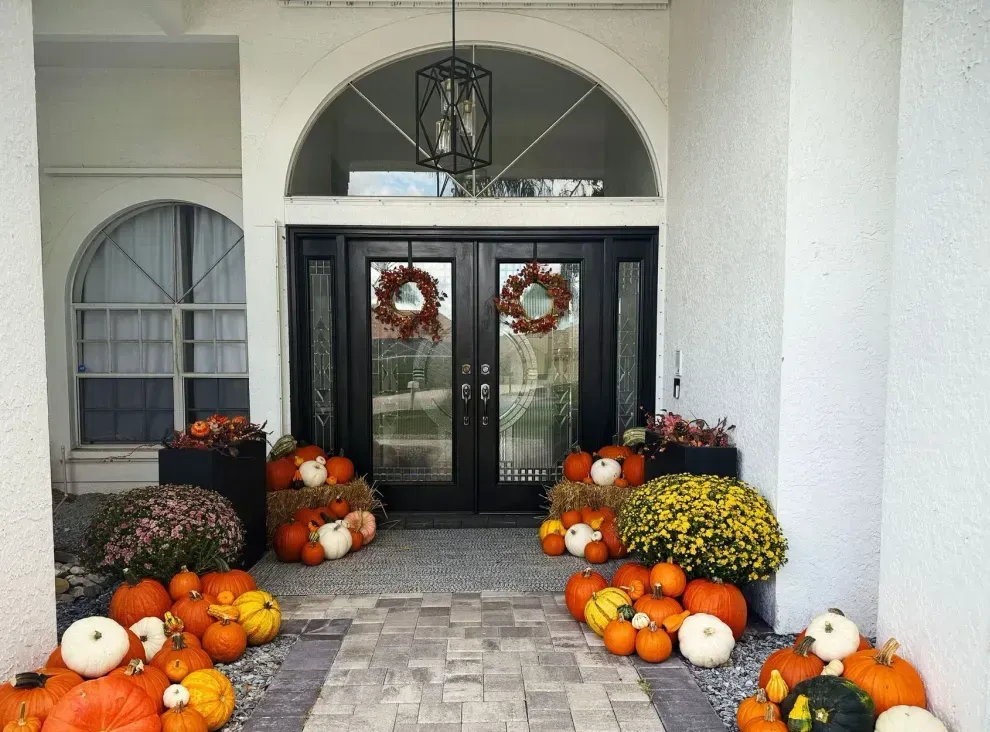 Fall-decorated porch with pumpkins, mums, and wreaths on the door. Black double doors under an arched entryway.