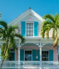 Pink and white two-story house with turquoise shutters and balcony, framed by palm trees against a blue sky.