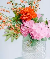 Vase of bright flowers: orange, red, and pink blooms with green foliage. Set on a white table.