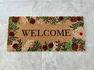 Welcome mat with brown text and a decorative pinecone border on a gray tiled floor.