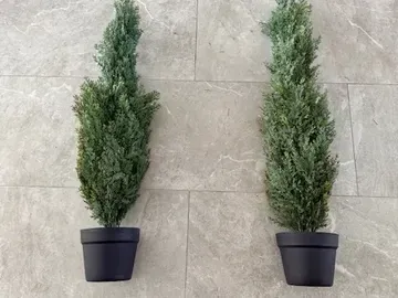 Two artificial cypress trees in black pots on a gray tile surface.