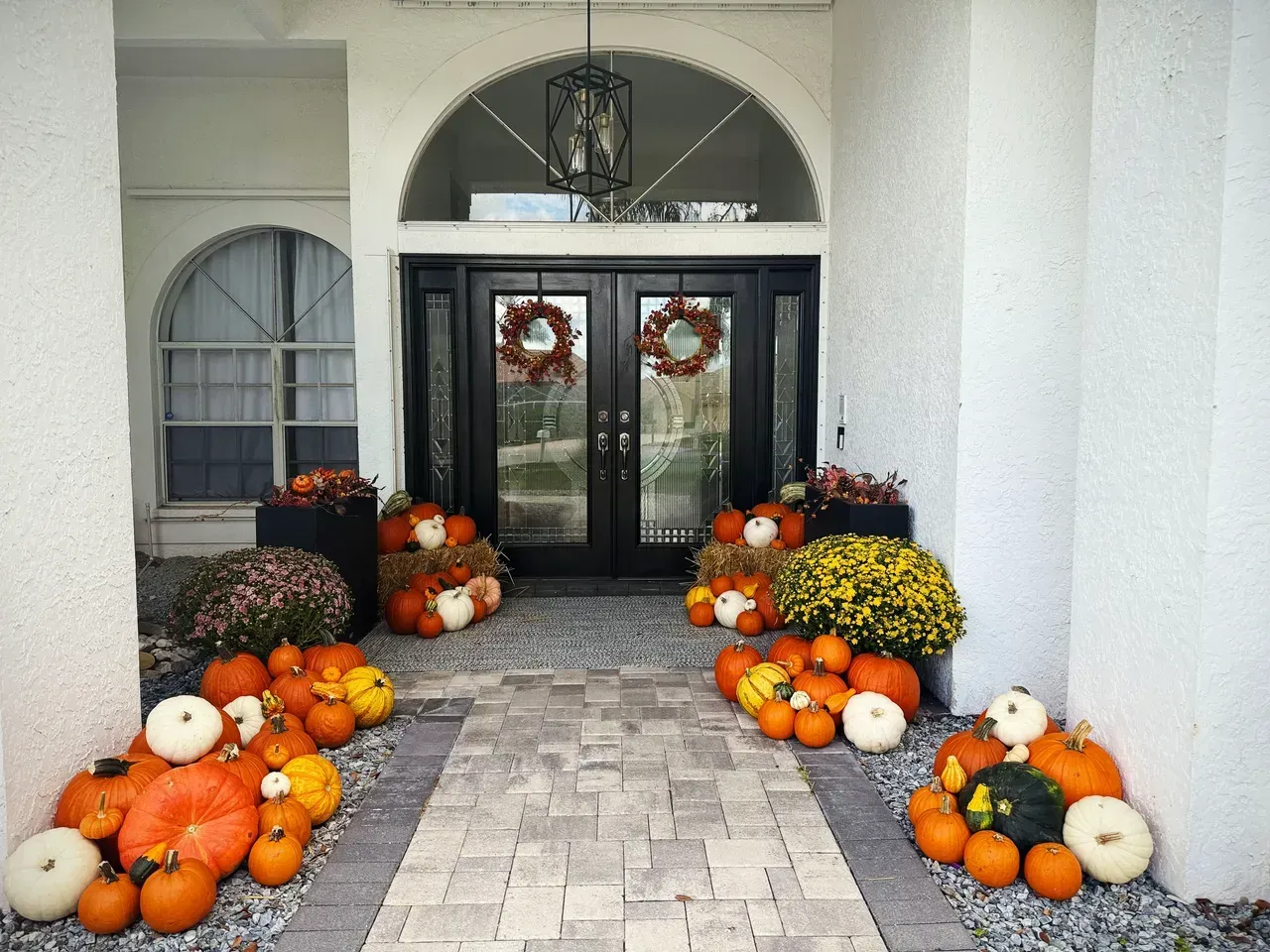 Fall porch decorated with pumpkins, mums, and wreaths; dark double doors.