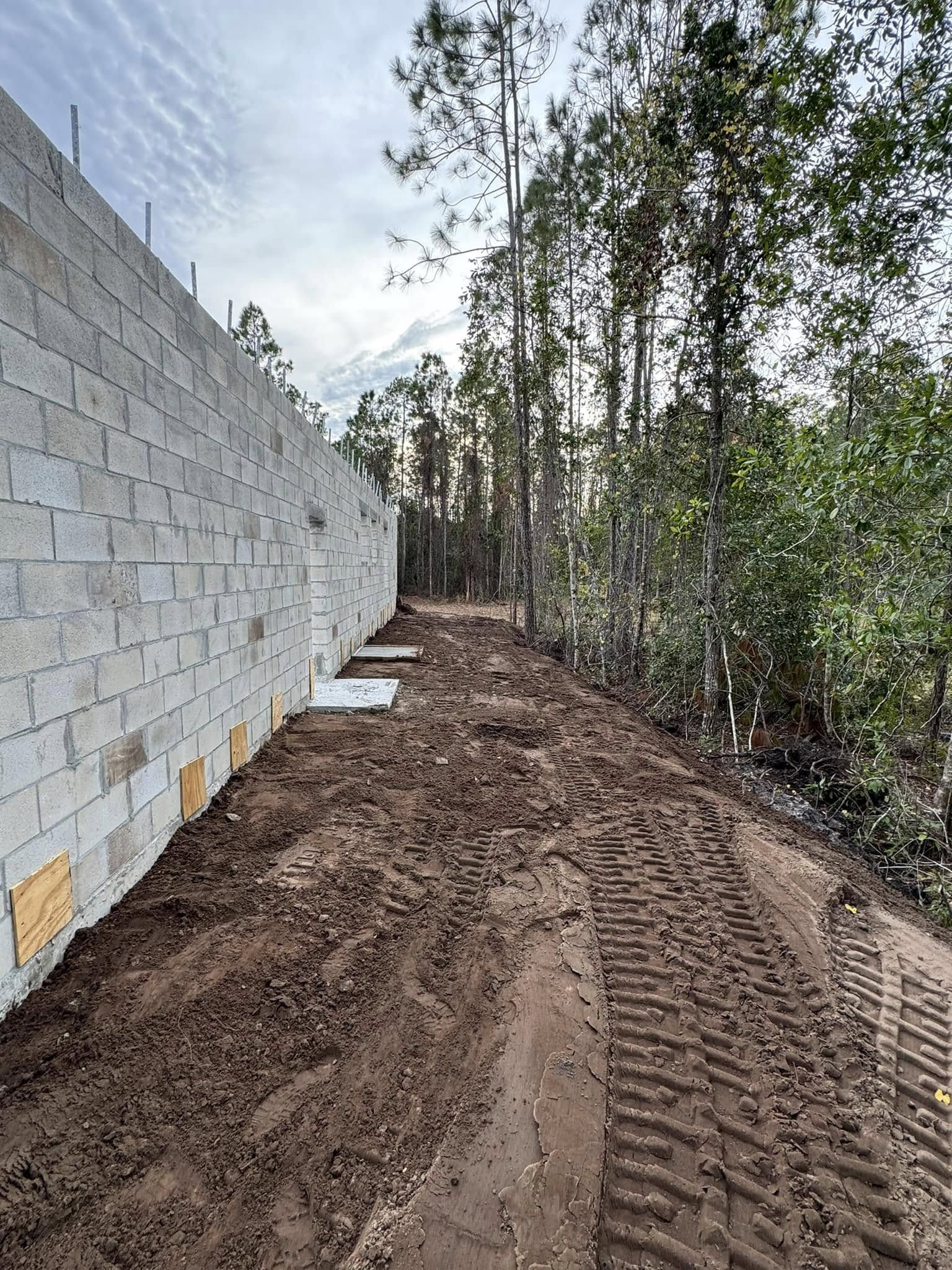 A cinder block wall borders a muddy path with tractor tracks, beside a wooded area under a cloudy sky.