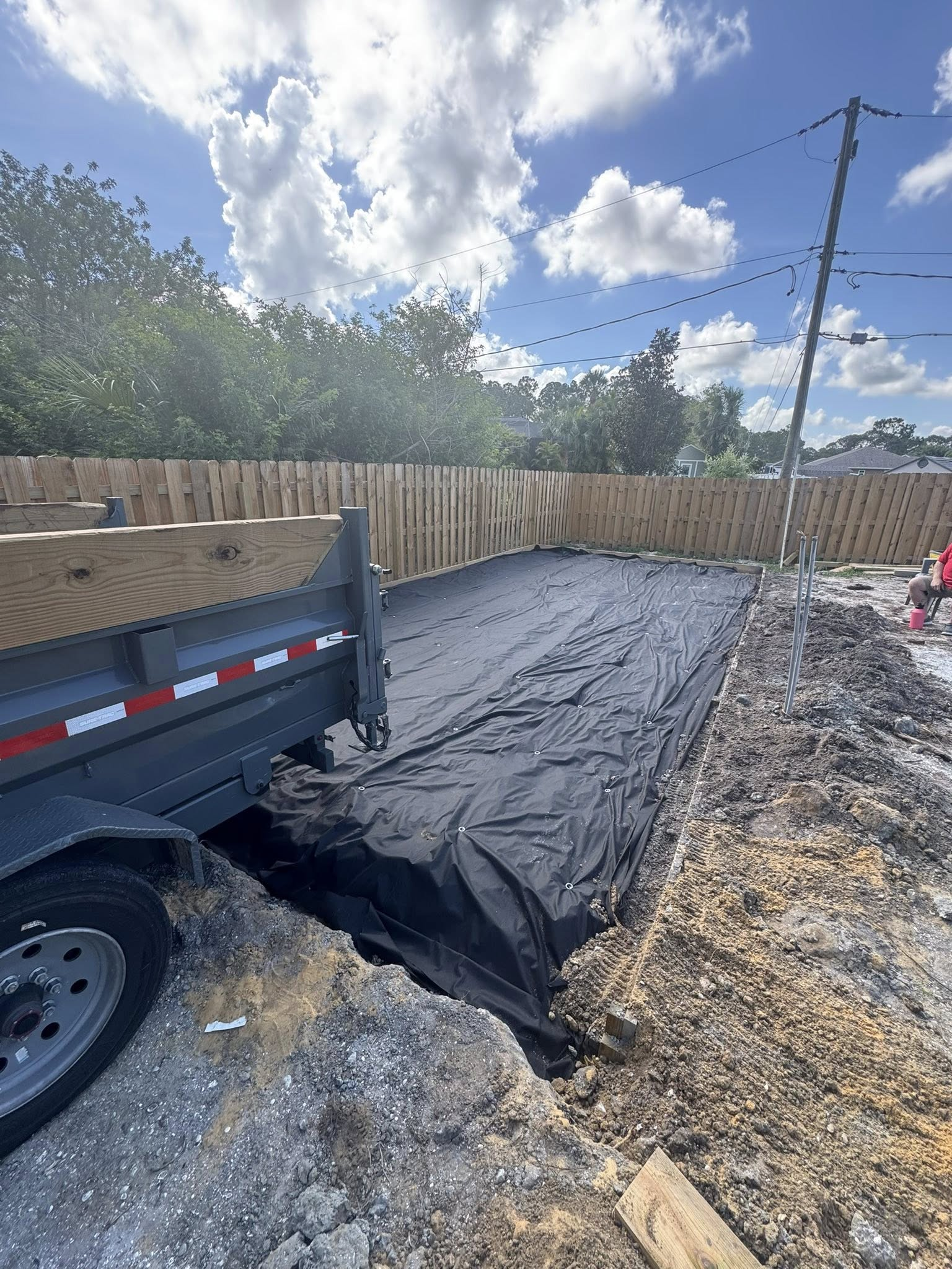 Dump truck beside a dug out area covered in black fabric, near a wooden fence and power pole.