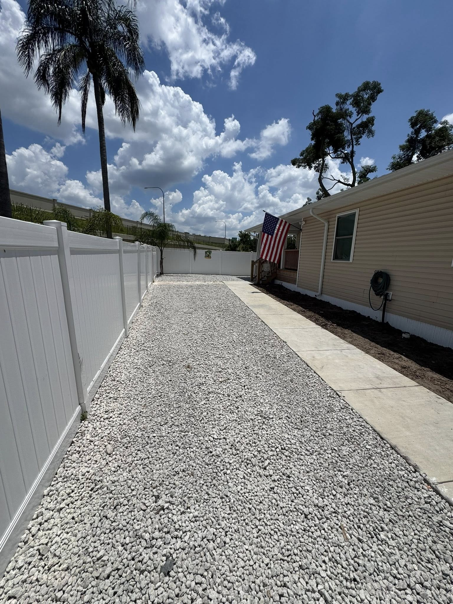 Gravel pathway beside a tan building and white fence, American flag hanging, sunny day.