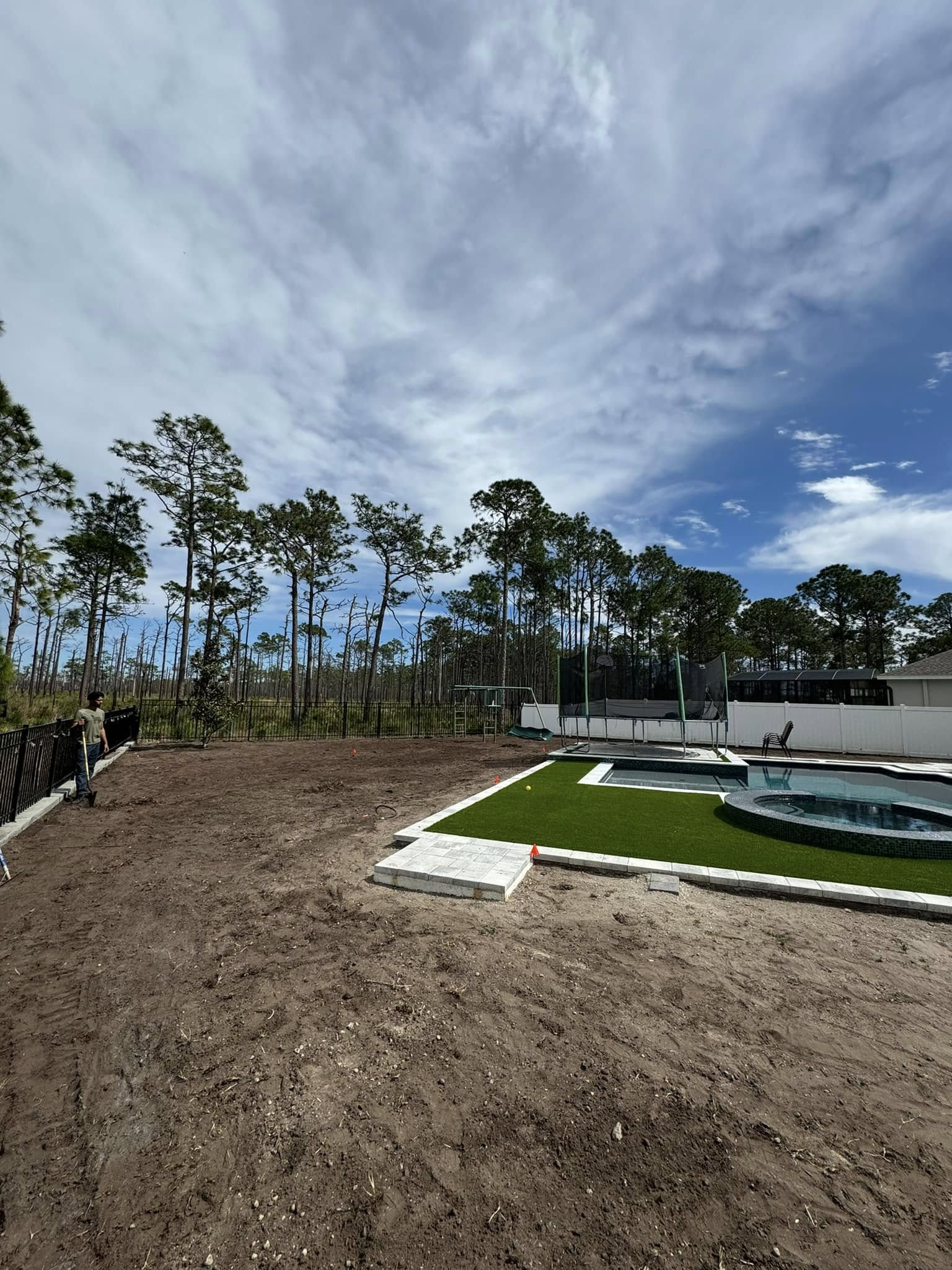 Backyard with pool, trees, and cloudy sky. Unfinished lawn, person near fence.