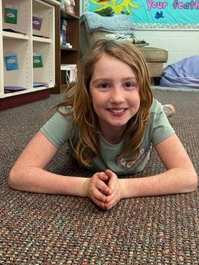 A young girl with long hair wearing a green t-shirt lying on her stomach on the floor smiling for the camera 