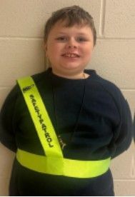A young boy wearing a black sweatshirt and a safety patrol badge smiling for the camera