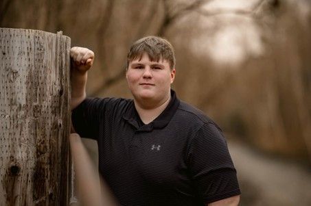 A young man wearing a black collared short sleeve shirt leaning against a wooden log
