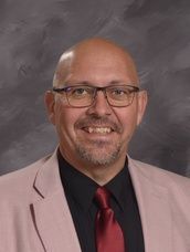 Man wearing glasses, a pink suite, black shirt, and burgundy tie smiling 