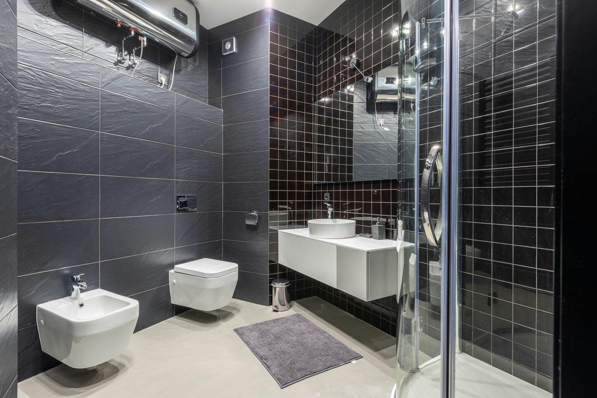 Modern black-tiled bathroom with toilet, bidet, sink, mirror, and glass shower stall