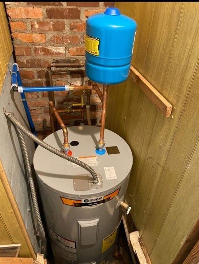 A water heater is sitting in a corner of a room next to a brick wall.