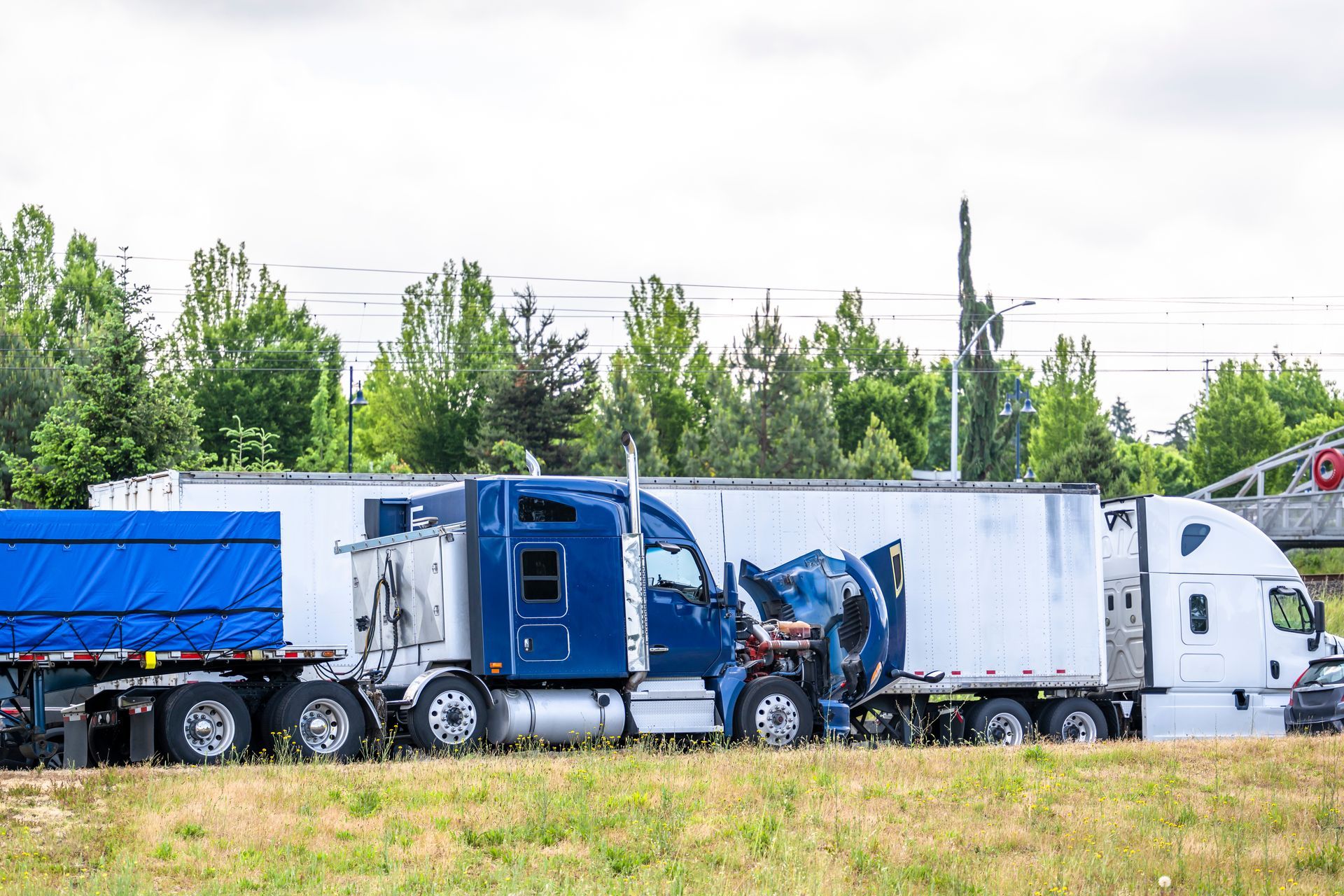 Damaged blue semi-truck cab with crumpled front, sandwiched between two trailers, roadside setting.