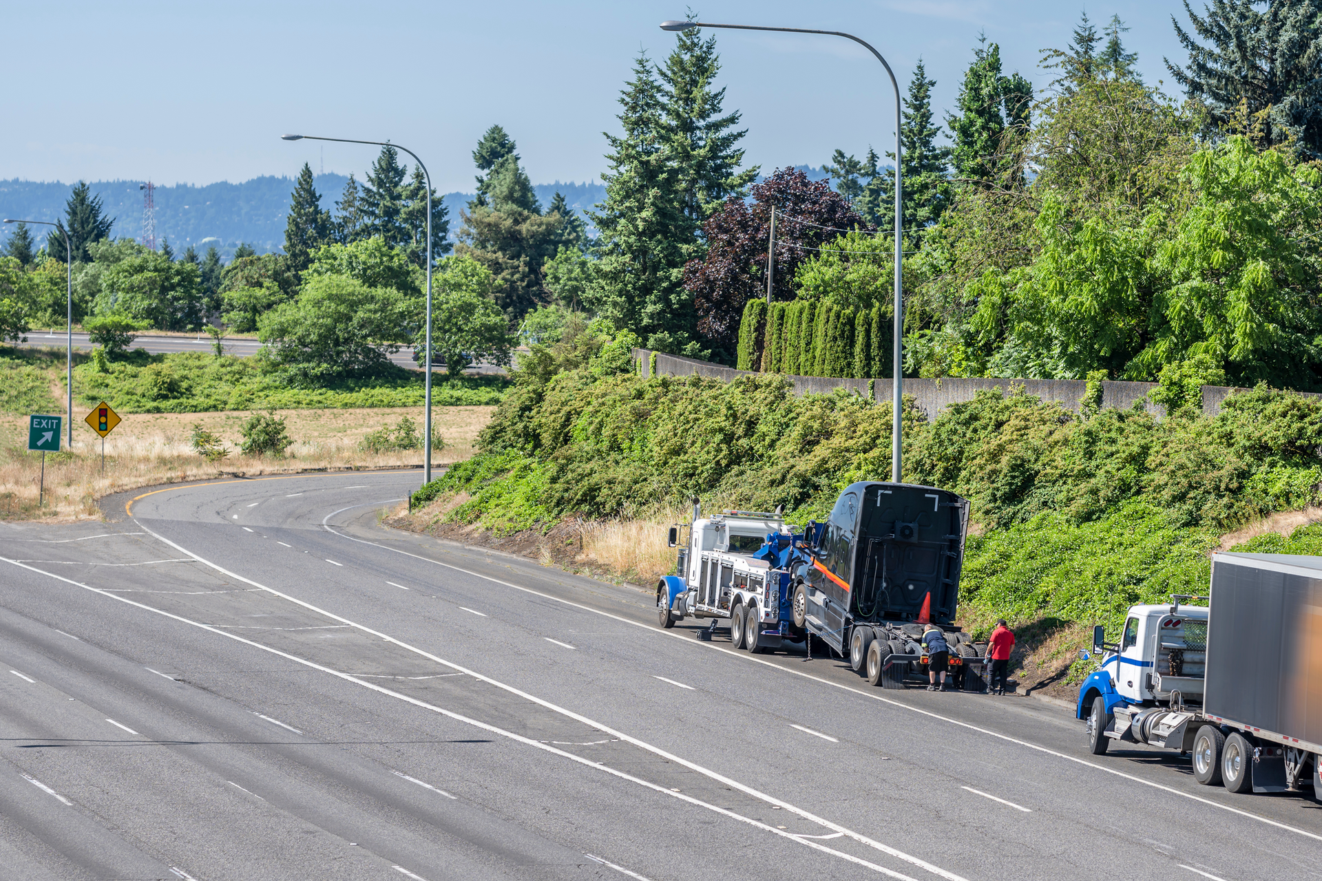 Two semi-trucks parked on a highway shoulder next to a grassy hill. One person is beside the truck.