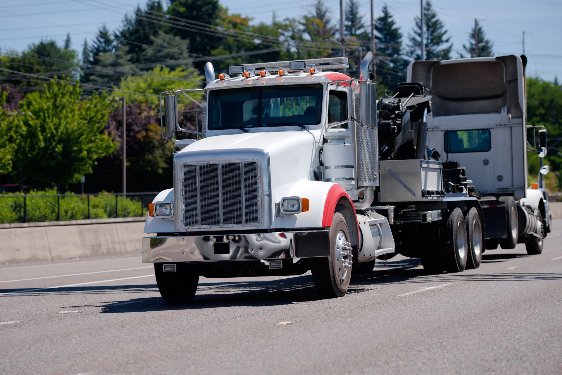 White semi-truck with red fenders on asphalt road, trees and blue sky background.